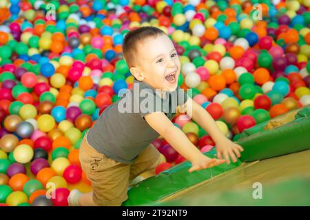 Joyeux garçon souriant grimpe sur un toboggan dans une chambre d'enfants ou un centre de divertissement pour enfants. Boules multicolores. Heureux passe-temps actif et Banque D'Images