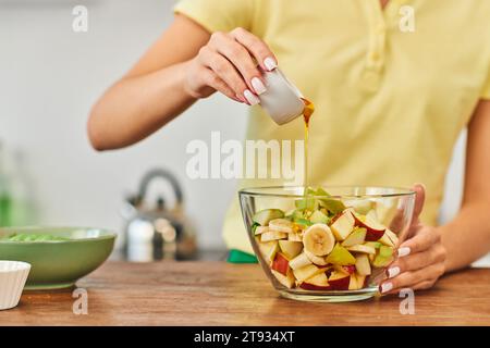 vue recadrée de femme versant du miel dans un bol avec des fruits hachés, salade végétarienne saine Banque D'Images