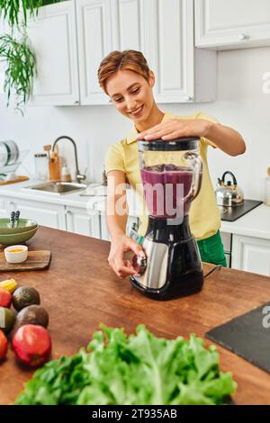 femme végétarienne enchantée broyant des fruits frais dans un mélangeur électrique, délicieuse recette de smoothie Banque D'Images