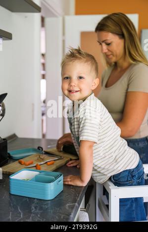 Vue latérale de la jeune mère en vêtements décontractés regardant vers le bas avec le fils souriant regardant la caméra tout en se tenant ensemble dans la cuisine et préparant la boîte à lunch Banque D'Images