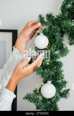 Main de femme caucasienne méconnaissable décorant la guirlande de noël en tenue élégante, mettant l'ornement blanc sur l'arbre, tenant une orna de noël Banque D'Images