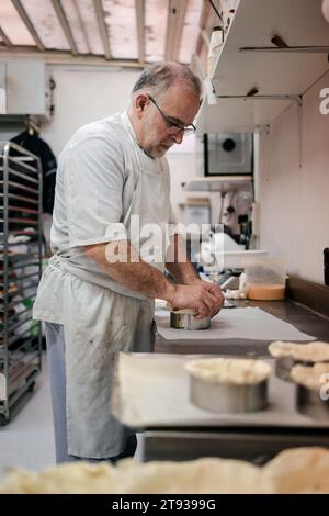 Chef pâtissier senior avec tablier blanc et verres faisant un gâteau dans la cuisine d'une pâtisserie Banque D'Images