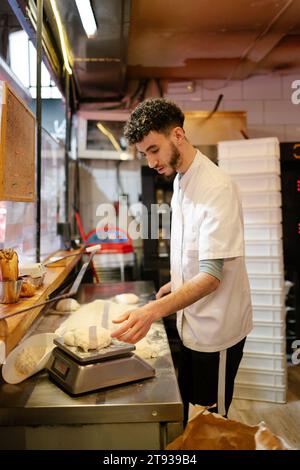Jeune homme de pizza arabe en tablier blanc pesant de la pâte sur une balance dans la cuisine d'une pizzeria Banque D'Images