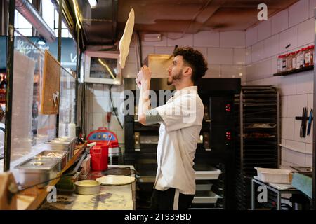 Portrait d'un jeune homme de pizza arabe en tablier blanc jetant la pâte en l'air dans une pizzeria Banque D'Images