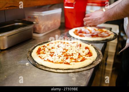 Portrait d'une pizza à la tomate et au fromage en cours de préparation avant d'entrer dans le four avec les mains de l'homme de pizza en arrière-plan préparant une autre pizza Banque D'Images