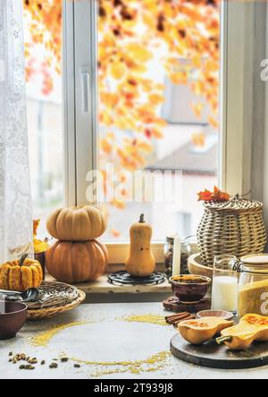 Nature morte de cuisine d'automne avec diverses citrouilles, ustensiles de cuisine et ingrédients à la table en béton avec fond de fenêtre avec feuillage d'automne Banque D'Images