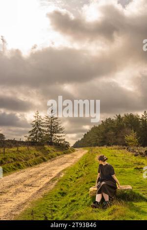 Fille en robe assis sur un rocher de grès regardant loin de la caméra le long d'une piste boueuse et humide à Osmotherly, North Yorkshire, Angleterre, Royaume-Uni Banque D'Images