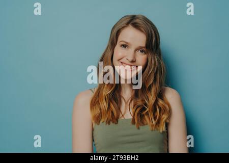 Femme radieuse avec des cheveux ondulés fluides et débardeur, partageant un sourire lumineux, placé sur un fond sarcelle. Banque D'Images