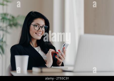 Femme d'affaires souriante utilisant smartphone à son espace de travail avec ordinateur portable et tasse à café en arrière-plan Banque D'Images