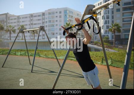 Travailler dur. Vue de côté d'un jeune homme sportif caucasien déterminé avec un corps parfait en vêtements de sport, faisant des pompes avec des sangles de fitness à l'extérieur en urb Banque D'Images