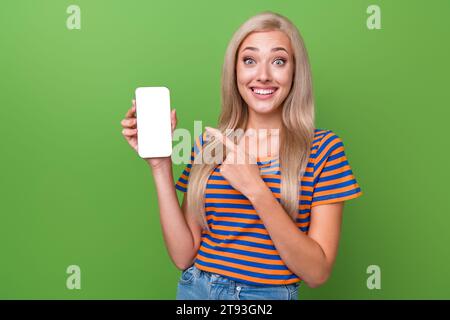 Photo de jeune femme excitée porter t-shirt rayé indiquant smartphone nouvelle mise à jour tiktok pour les utilisateurs isolés sur fond de couleur verte Banque D'Images