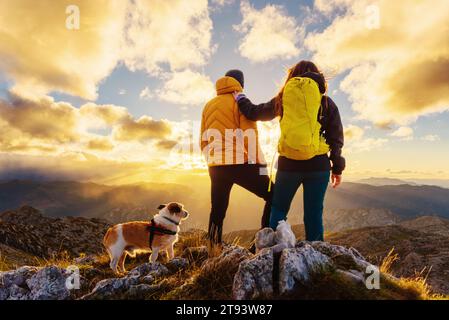 un couple de randonneurs avec leur chien regardant le coucher du soleil ensemble du sommet d'une montagne. sports et activités de montagne et de plein air. Banque D'Images