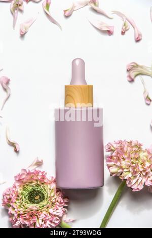 Bouteille de maquillage violet pastel avec pipette sur fond blanc avec fleurs roses , vue de dessus. Produit de beauté moderne pour les soins de la peau avec espace de copie. Banque D'Images