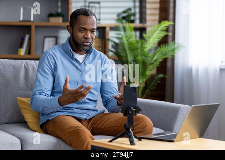 Homme concentré sérieux enregistrement vidéo en ligne, homme afro-américain utilisant le téléphone sur trépied assis sur le canapé dans le salon. Banque D'Images