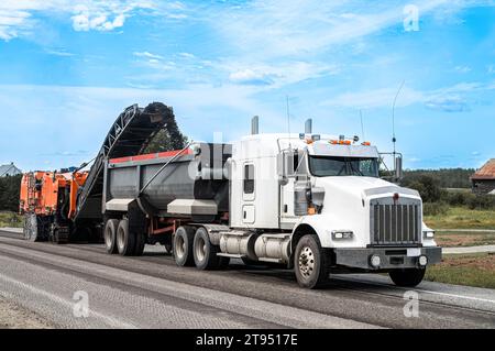 Rabotage orange enlevant une couche d'asphalte d'une route et la transférant à un camion semi-remorque blanc Banque D'Images