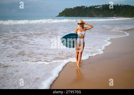 Femme athlétique en maillot de bain élégant tenant la planche de surf sur la plage tropicale, se préparer à surfer vagues, mode de vie actif et concept de sport Banque D'Images