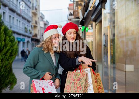 De jeunes amies hispaniques positives souriant et regardant loin tout en se tenant sur le trottoir avec des sacs à provisions et en pointant du doigt les produits exposés Banque D'Images