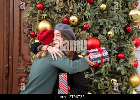 Vue latérale de souriants jeunes amies hispaniques embrassant les uns les autres tout en se tenant près de l'arbre de Noël décoré et échangeant des cadeaux à la lumière du jour Banque D'Images