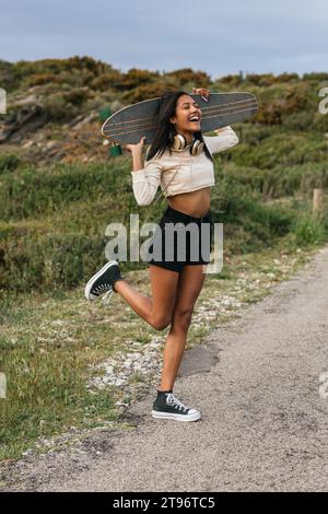 Corps complet de jeune femme gaie ethnique debout sur une jambe avec une longue planche derrière la tête et un casque sur le cou et rire avec la bouche ouverte Banque D'Images