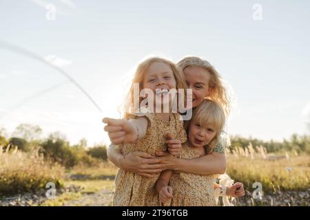 Heureuse jeune femme aux cheveux blonds embrassant de mignonnes petites filles portant des robes assorties et regardant la caméra contre la lumière du soleil dans la campagne Banque D'Images