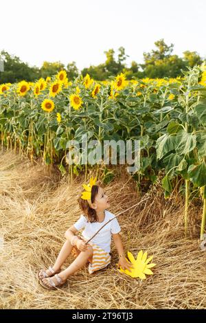 Petite fille avec la bouche ouverte assis dans la prairie de tournesol en fleurs dans la campagne et regardant vers le haut tout en tenant la branche d'arbre et profiter de l'été Banque D'Images