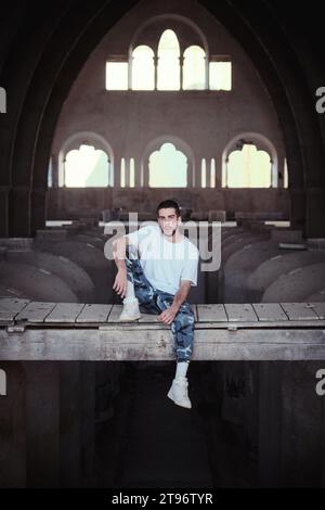 Corps entier de mâle calme regardant la caméra tout en étant assis sur le passage supérieur de l'ancienne installation de la cathédrale négligée avec fenêtre Banque D'Images