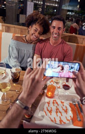 D'en haut d'un mâle méconnaissable prenant la photo d'heureux jeune couple diversifié assis à table tout en dînant dans le pub Banque D'Images
