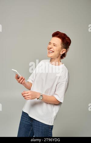 personne queer roux joyée en t-shirt blanc avec smartphone riant avec les yeux fermés sur le gris Banque D'Images