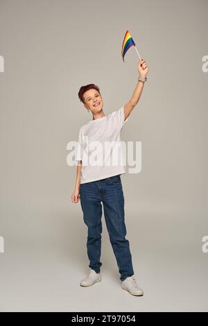 Joyeuse personne queer en t-shirt blanc tenant un petit drapeau LGBT à la main levée tout en se tenant debout sur le gris Banque D'Images