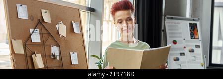 souriant queer personne regardant des documents près du panneau de liège avec des notes de papier dans le bureau, bannière Banque D'Images