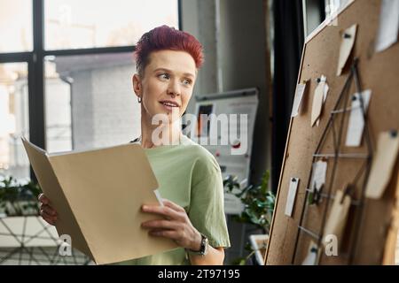 personne souriante queer avec des documents regardant le panneau de liège avec des notes de papier dans le bureau moderne Banque D'Images