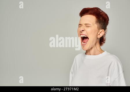 personne queer émotionnelle dans un t-shirt blanc debout et criant avec les yeux fermés sur fond gris Banque D'Images