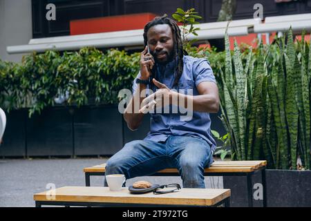 Homme africain avec des dreadlocks et barbe, assis à l'extérieur, discutant au téléphone, la technologie et le concept de modes de vie, copier l'espace. Banque D'Images
