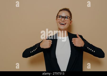 Femme professionnelle optimiste donnant les pouces vers le haut, rayonnant avec confiance dans des vêtements intelligents et des lunettes, isolé sur un fond beige Banque D'Images