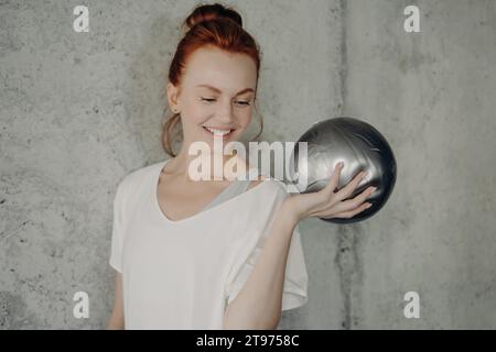 Joyeuse rousse dans une tenue décontractée pose de manière ludique avec une balle d'exercice argentée dans une salle de gym minimaliste Banque D'Images