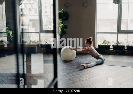 Une femme rousse s'étire avec une balle de Pilates dans un studio ensoleillé, incarnant bien-être et équilibre à chaque mouvement Banque D'Images