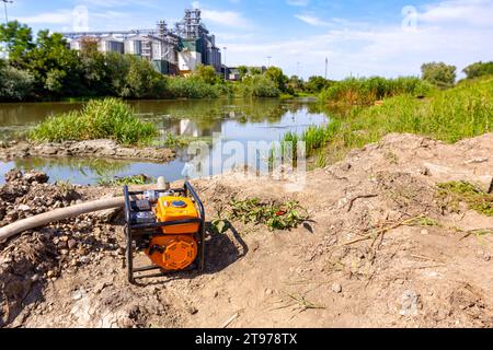 Pompe à moteur orange avec long tuyau pour remplir le réservoir avec de l'eau placée sur la côte, la construction du pont est en cours, à travers la rivière sont de grands silos à grain Banque D'Images