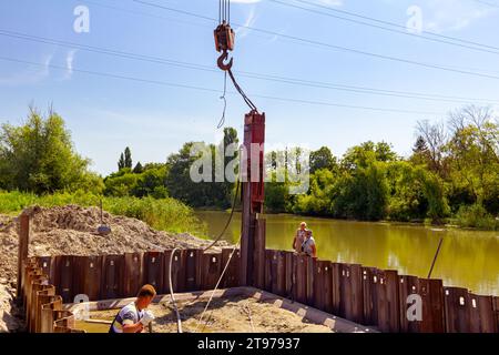 Le camion de grue mobile avec marteau pneumatique installe des pieux métalliques dans la fondation de pont le long de la rive de la rivière sur le chantier de construction. Le travailleur gère avec l'arc Banque D'Images