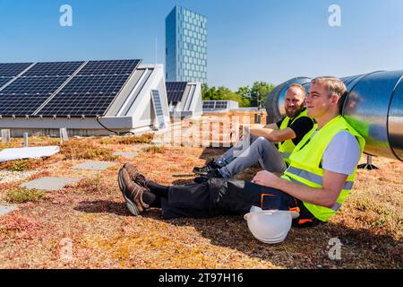 Deux techniciens en panneaux solaires prenant une pause bien méritée sur le toit d’un immeuble corporatif Banque D'Images