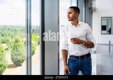Homme d'affaires occasionnel regardant par la fenêtre tenant une tasse de café Banque D'Images