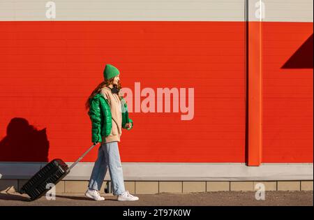 Jeune femme portant un chapeau tricoté marchant avec une valise à roulettes devant le mur rouge Banque D'Images