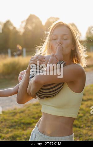 Mère aimante embrassant le pied de bébé dans le parc au coucher du soleil Banque D'Images