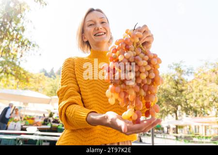 Femme heureuse tenant bouquet de raisins frais au marché fermier Banque D'Images