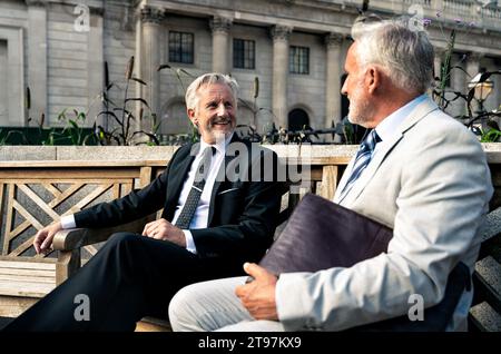 Souriant hommes d'affaires ayant des discussions ensemble sur le banc en face de la construction financière Banque D'Images