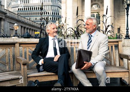 Heureux hommes d'affaires seniors ayant des discussions ensemble sur le banc devant les bâtiments Banque D'Images