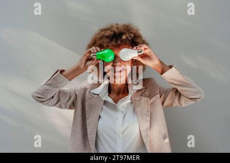 Heureuse jeune femme d'affaires tenant des ampoules au-dessus des yeux devant le mur Banque D'Images