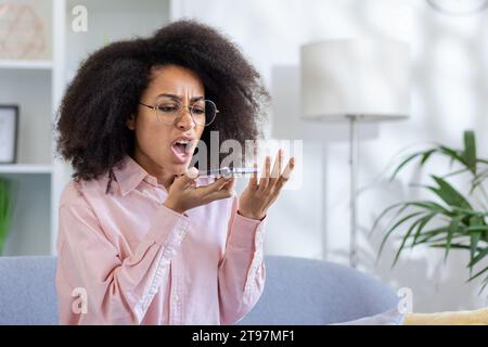 Jeune femme afro-américaine en colère assise sur le canapé à la maison et parlant émotionnellement au téléphone à travers le haut-parleur. Banque D'Images