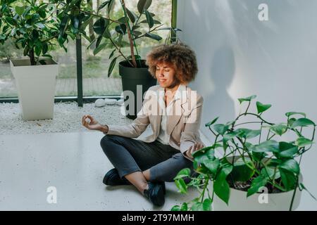 Femme d'affaires souriante pratiquant la méditation près des plantes dans le bureau Banque D'Images