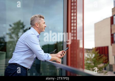 Homme d'affaires réfléchi appuyé sur la rampe avec téléphone portable devant le verre Banque D'Images