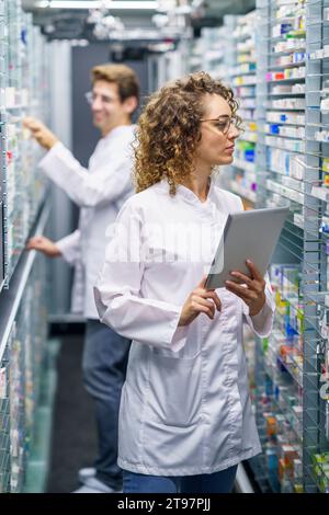 Pharmacien tenant le PC comprimé et examinant les médicaments sur les étagères de la pharmacie Banque D'Images